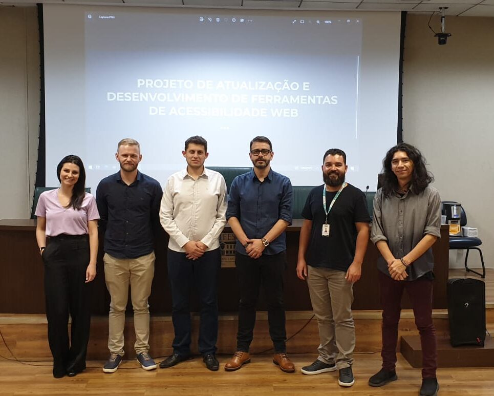 Integrantes do projeto, uma mulher e cinco homens, em p&eacute;, em frente ao palco. No fundo, tel&atilde;o com um slide projetado, com o t&iacute;tulo do projeto Atualiza&ccedil;&atilde;o e desenvolvimento de ferramentas para acessibilidade web
