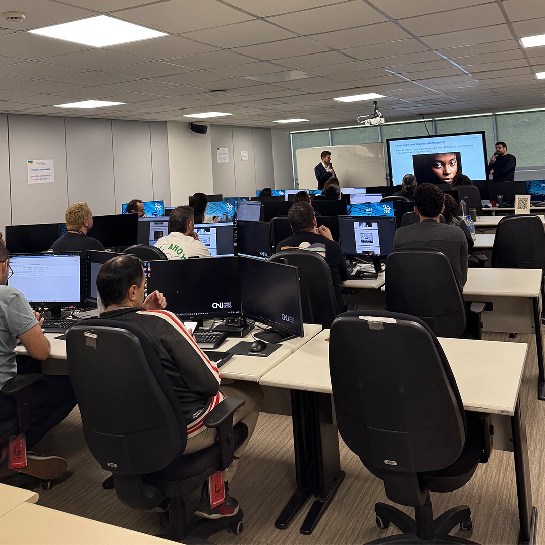 Sala com pessoas sentadas em frente a computadores e pessoa em pé apresentando conteúdo.