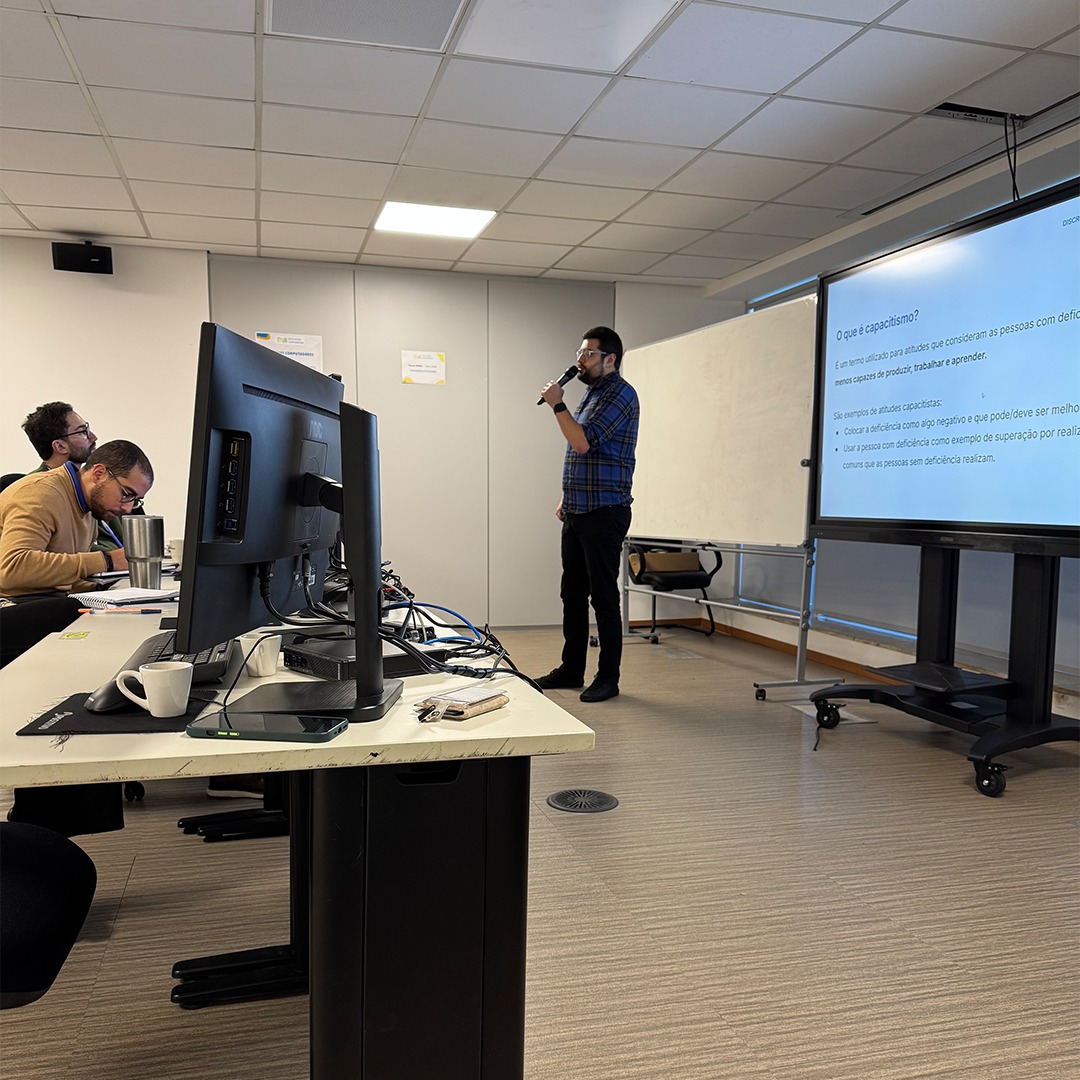Pessoa em pé ao lado de tela com slide; outras estão sentadas em frente a computadores.