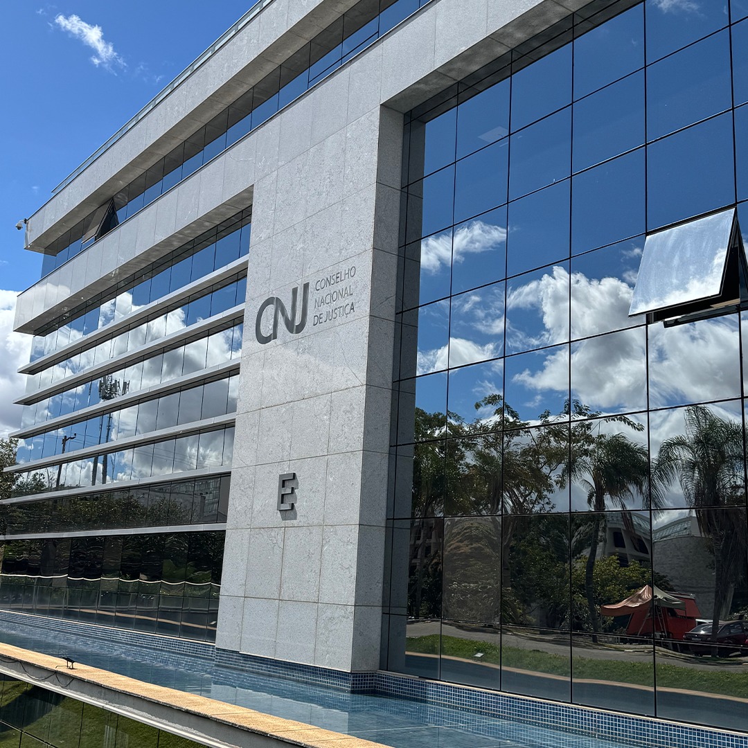 Fachada do pr&eacute;dio do Conselho Nacional de Justi&ccedil;a (CNJ), com paredes espelhadas refletindo c&eacute;u, nuvens e &aacute;rvores ao redor.